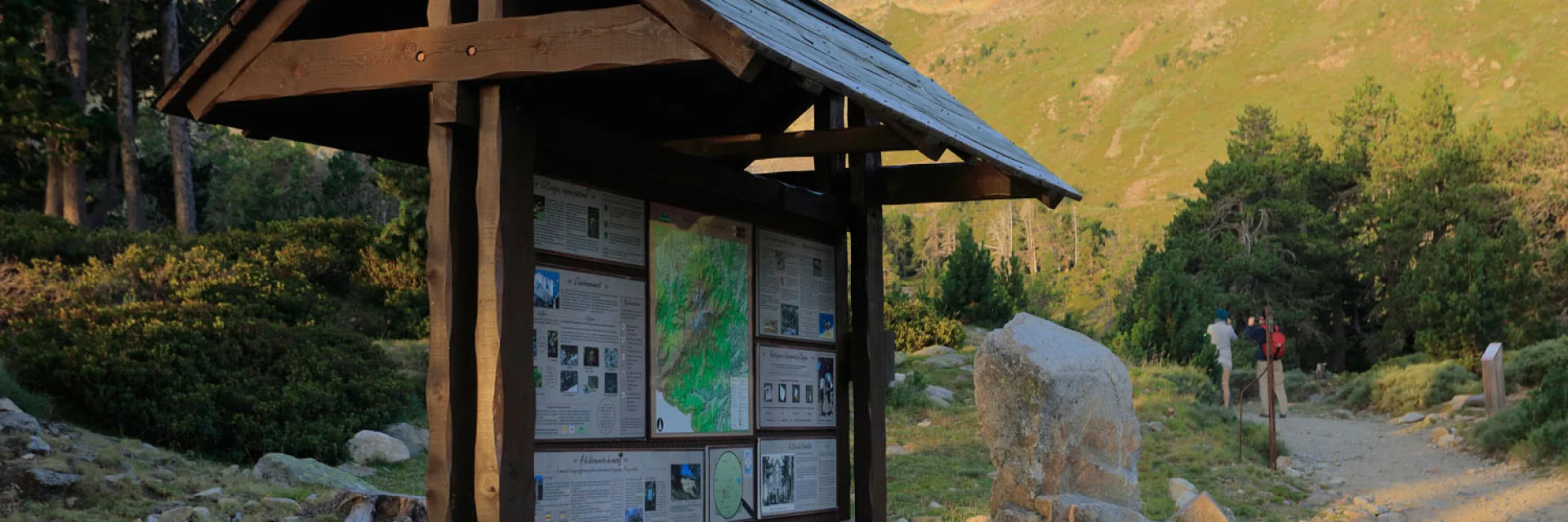 Panneau d’information en bois près d’un sentier pierreux en montagne, entouré d’arbres et de pentes herbeuses.