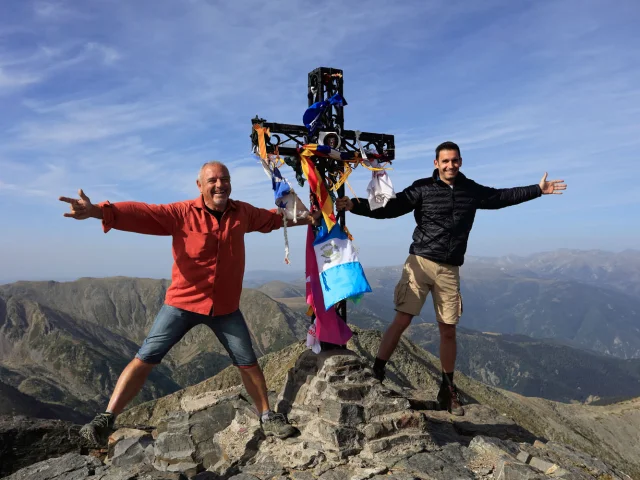 Personnes au sommet d’une montagne, bras ouverts devant une grande croix ornée de rubans colorés.