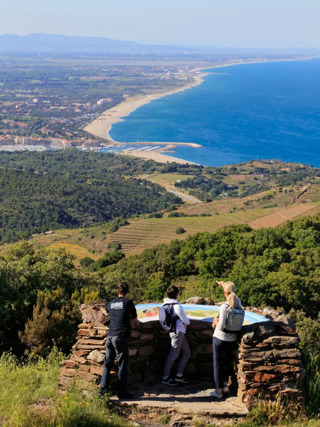 Trois personnes observent un panorama côtier depuis un point de vue en pierre, avec la mer et la plaine au loin