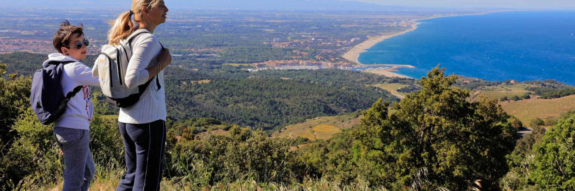 Deux personnes équipées de sacs à dos observent un large panorama côtier depuis un sentier en hauteur sous un ciel dégagé.