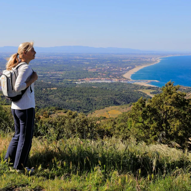 Deux personnes équipées de sacs à dos observent un large panorama côtier depuis un sentier en hauteur sous un ciel dégagé.