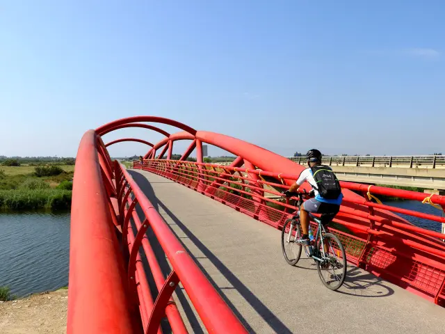 Cycliste traversant une passerelle rouge au-dessus d’un cours d’eau, dans un paysage ouvert.