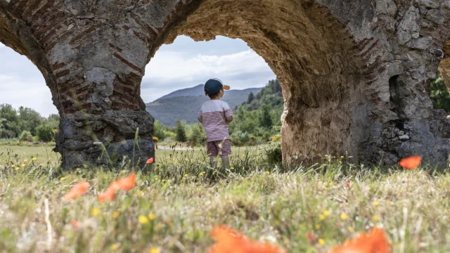 Enfant vu de dos, debout dans l’herbe devant une arche en pierre, avec des collines en arrière-plan.