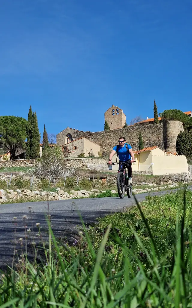 Cycliste roulant sur une route de campagne devant un village aux bâtiments en pierre et un ancien clocher, sous un ciel bleu dégagé.