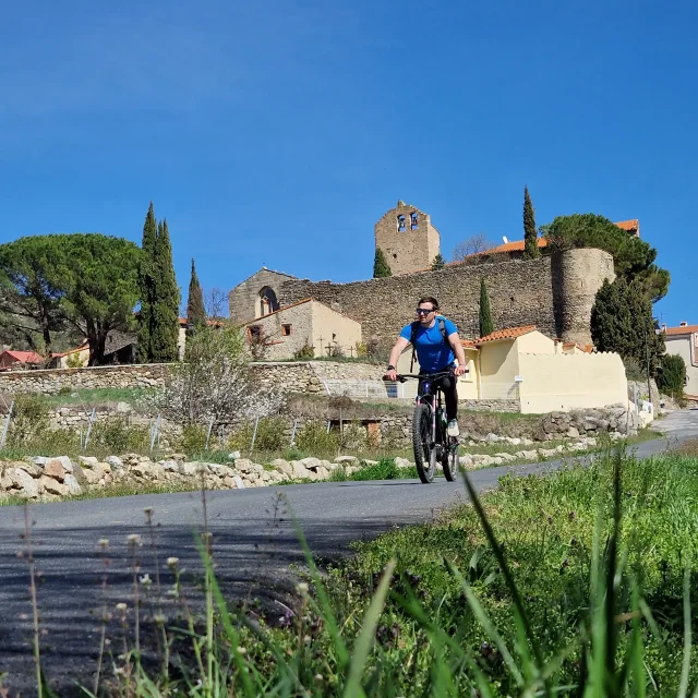 Cycliste roulant sur une route de campagne devant un village aux bâtiments en pierre et un ancien clocher, sous un ciel bleu dégagé.