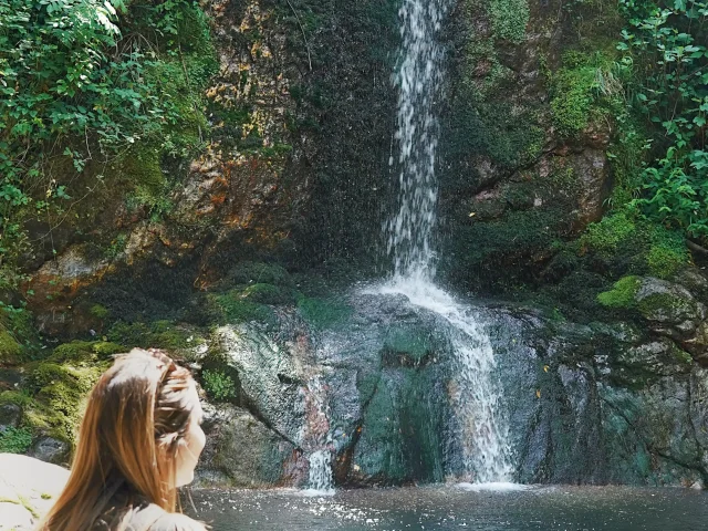 Personne de dos observant une chute d’eau se déverser le long d’une paroi rocheuse couverte de mousse, formant une vasque claire au pied de la cascade.