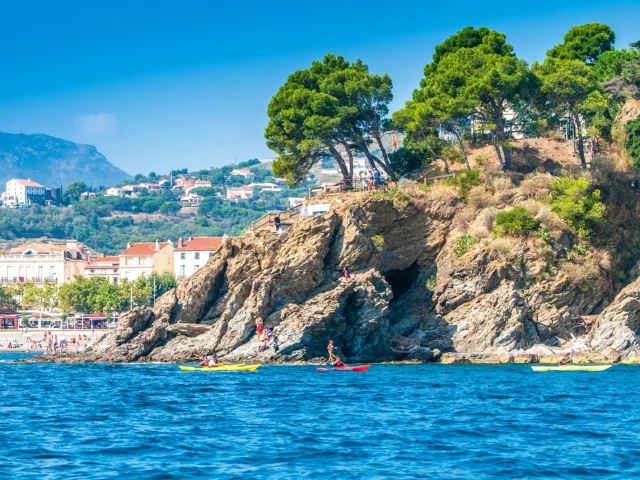 Kayaks naviguant près de rochers côtiers, avec une plage animée et des habitations en arrière-plan.