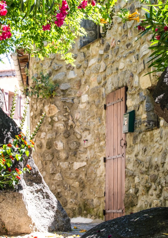 Ruelle en pierre bordée de gros rochers et de plantes fleuries, avec une porte en bois fermée sous une lumière ensoleillée.