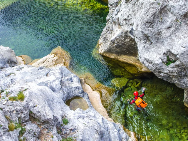 Personne équipée de casque et sac étanche avançant dans un bassin entre de larges rochers.