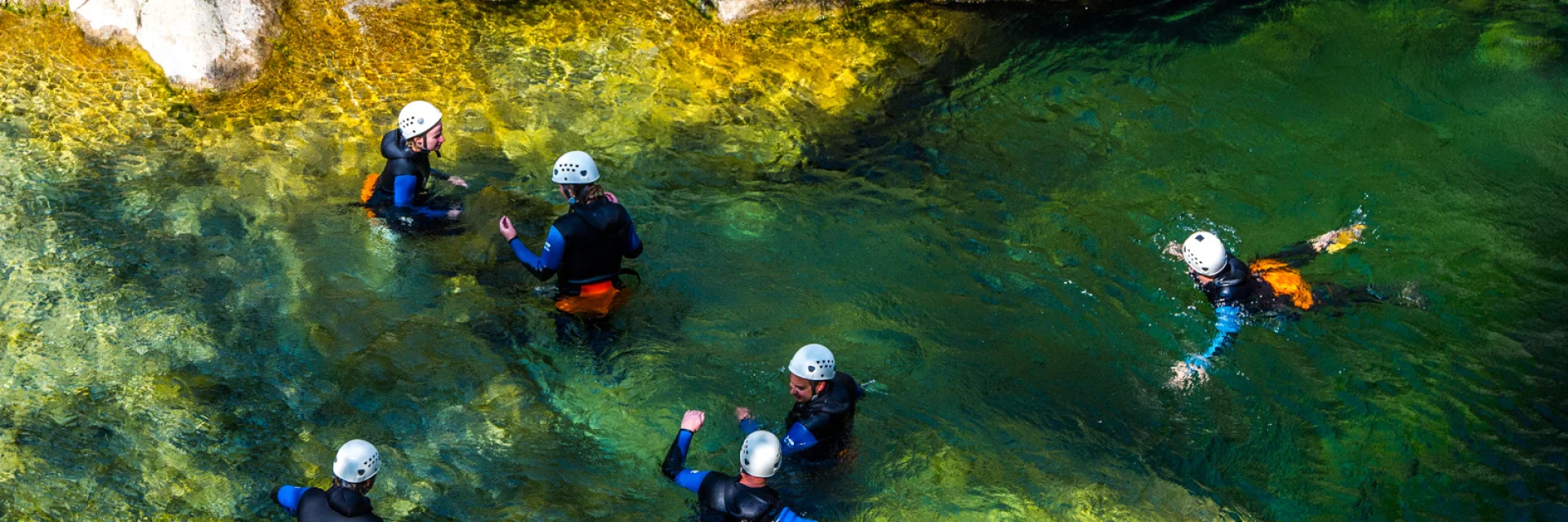 Groupe équipé de combinaisons et casques progressant dans un bassin d’eau claire au pied de rochers.
