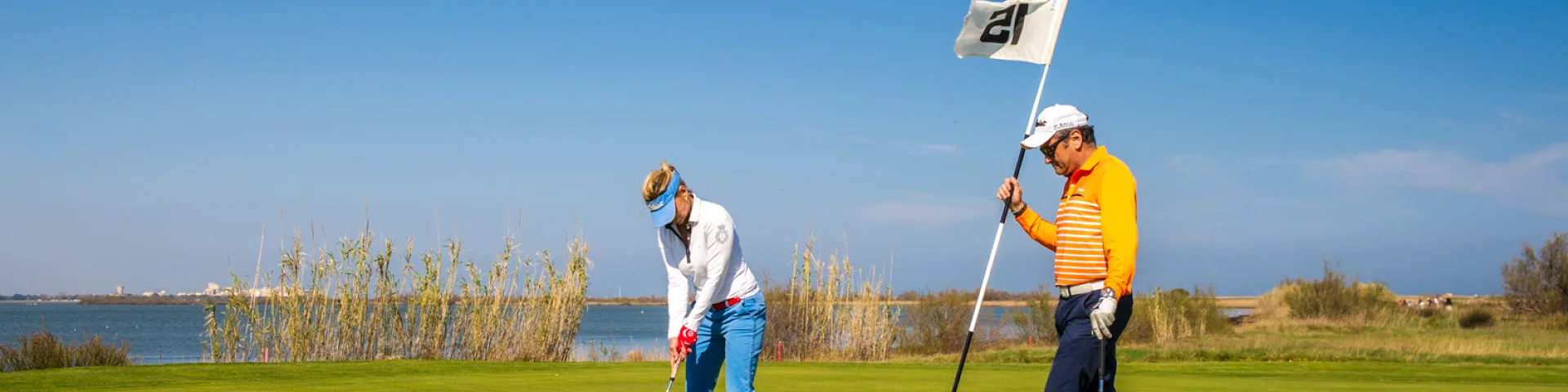 Deux golfeurs sur un green en bord d’étang, sous un ciel bleu dégagé, l’un prêt à putter, l’autre tenant le drapeau.