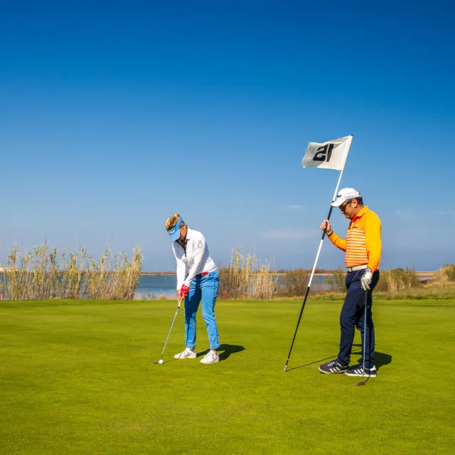Deux golfeurs sur un green en bord d’étang, sous un ciel bleu dégagé, l’un prêt à putter, l’autre tenant le drapeau.