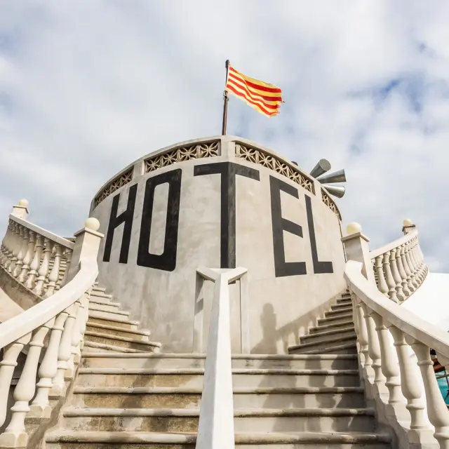 Escaliers symétriques menant à une façade circulaire portant l’inscription « Hôtel », sous un drapeau à rayures.