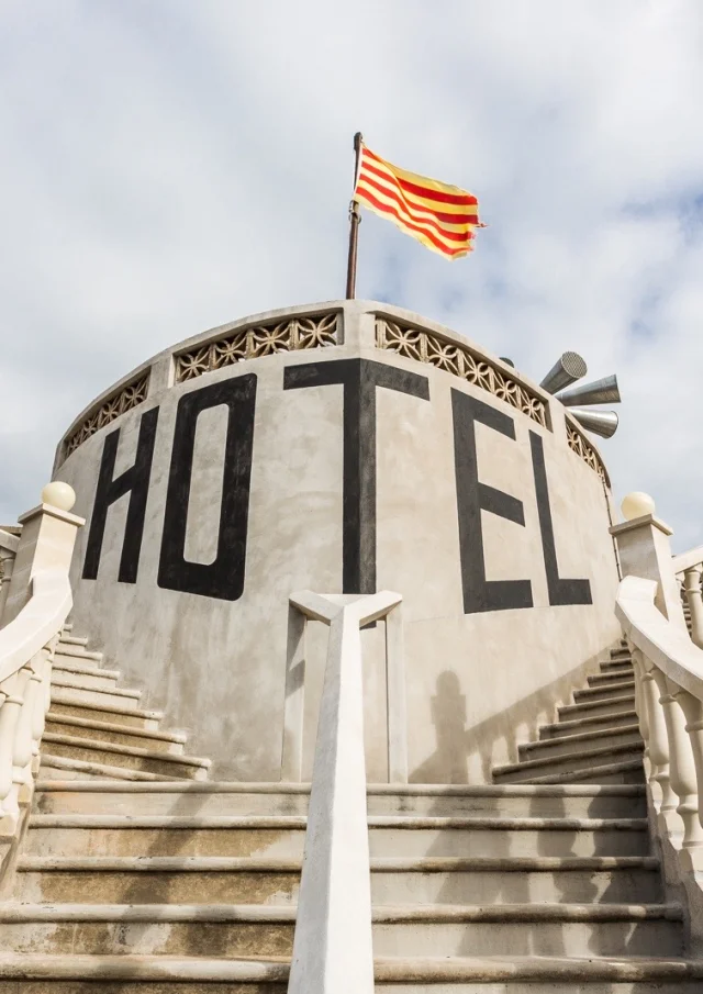 Escaliers symétriques menant à une façade circulaire portant l’inscription « Hôtel », sous un drapeau à rayures.