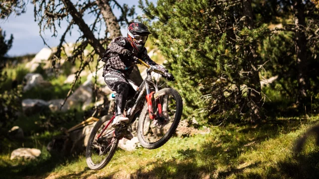 Cycliste équipé en descente VTT effectuant un saut sur une piste forestière.