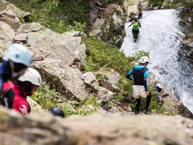 Groupe équipé de combinaisons et casques descendant un sentier rocheux vers un cours d’eau.