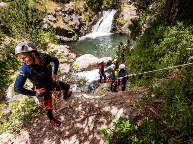 Groupe équipé de combinaisons et casques progressant sur un rocher près d’une cascade et d’un bassin naturel.