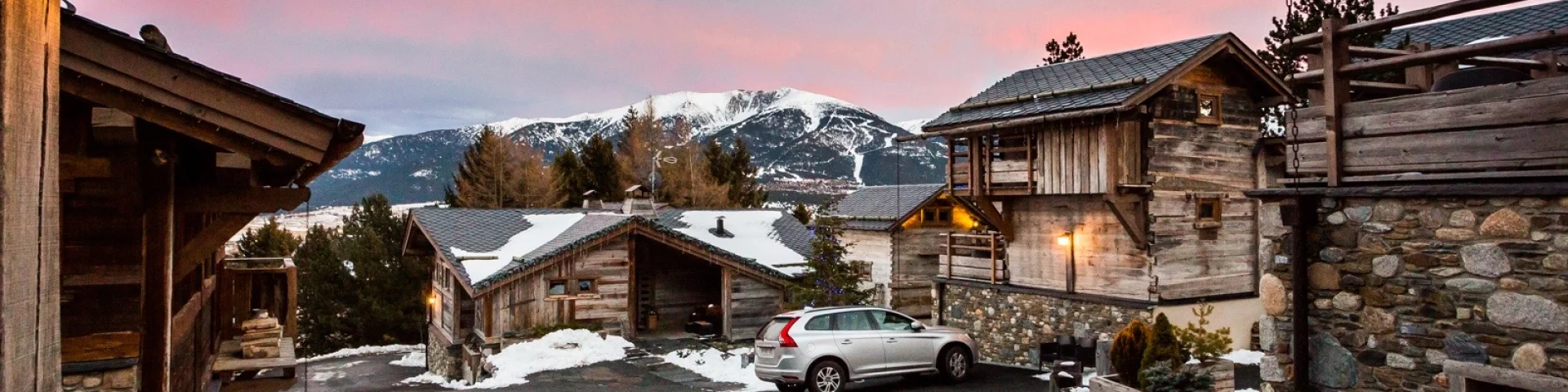 Vue en fin de journée sur les Chalets Secrets à Bolquère, avec un ciel rosé, des toits enneigés et les sommets du Canigó, du Gallinàs et du Cambre d’Aze en arrière-plan.