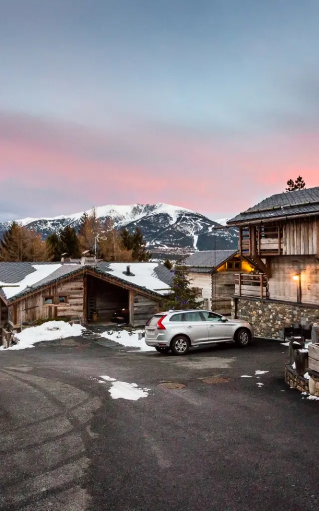Vue en fin de journée sur les Chalets Secrets à Bolquère, avec un ciel rosé, des toits enneigés et les sommets du Canigó, du Gallinàs et du Cambre d’Aze en arrière-plan.