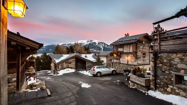 Vue en fin de journée sur les Chalets Secrets à Bolquère, avec un ciel rosé, des toits enneigés et les sommets du Canigó, du Gallinàs et du Cambre d’Aze en arrière-plan.