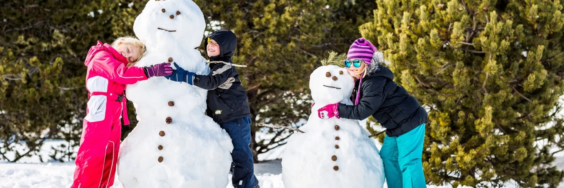 Trois enfants en tenue de ski sourient et posent devant deux grands bonshommes de neige qu’ils viennent de construire à La Calme, sur la neige blanche bordée de pins.