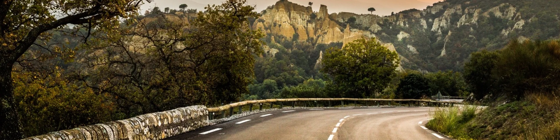 Route sinueuse bordée d’arbres menant vers les Orgues d’Ille-sur-Têt, formations rocheuses ocres se détachant sur fond de montagnes et du massif du Canigó au coucher du soleil.
