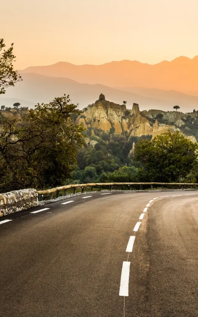 Route sinueuse bordée d’arbres menant vers les Orgues d’Ille-sur-Têt, formations rocheuses ocres se détachant sur fond de montagnes et du massif du Canigó au coucher du soleil.
