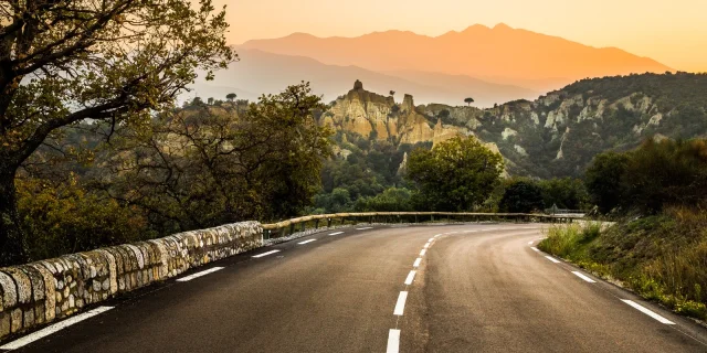 Route sinueuse bordée d’arbres menant vers les Orgues d’Ille-sur-Têt, formations rocheuses ocres se détachant sur fond de montagnes et du massif du Canigó au coucher du soleil.