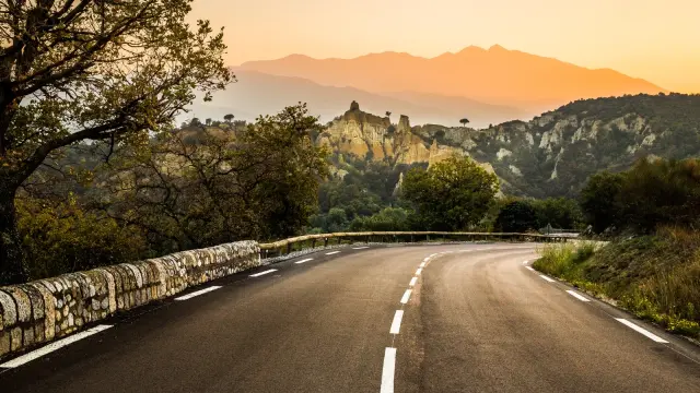 Route sinueuse bordée d’arbres menant vers les Orgues d’Ille-sur-Têt, formations rocheuses ocres se détachant sur fond de montagnes et du massif du Canigó au coucher du soleil.