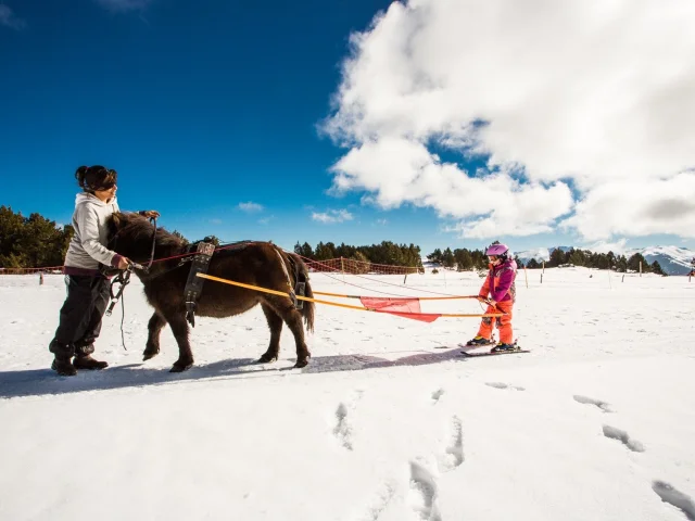 Enfant en tenue de ski tiré par un poney dans la neige, accompagné d’une adulte, sous un ciel bleu vif à La Calme, dans la station de Font-Romeu.