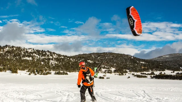 Personne pratiquant le snowkite sur l’aérodrome enneigé de La Quillane, tirée par une voile rouge et noire sous un ciel bleu, avec les montagnes du massif du Madres en arrière-plan.