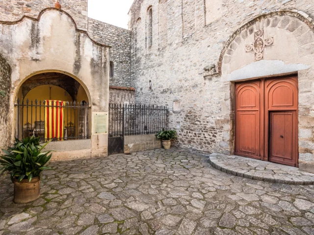 Cour intérieure de l’abbaye Sainte-Marie d’Arles-sur-Tech avec le portail en bois de l’église, une chapelle latérale protégée par une grille et ornée du drapeau catalan.