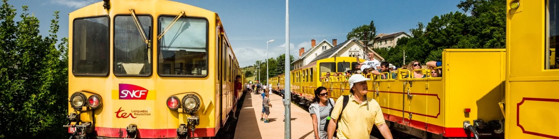 Le Train Jaune à quai en gare de Mont-Louis, avec des voyageurs descendant du train ou montant à bord. Les wagons jaunes emblématiques sont visibles sous un ciel bleu dégagé.