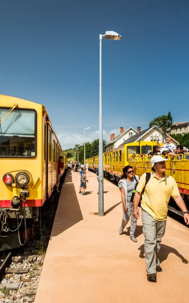 Le Train Jaune à quai en gare de Mont-Louis, avec des voyageurs descendant du train ou montant à bord. Les wagons jaunes emblématiques sont visibles sous un ciel bleu dégagé.