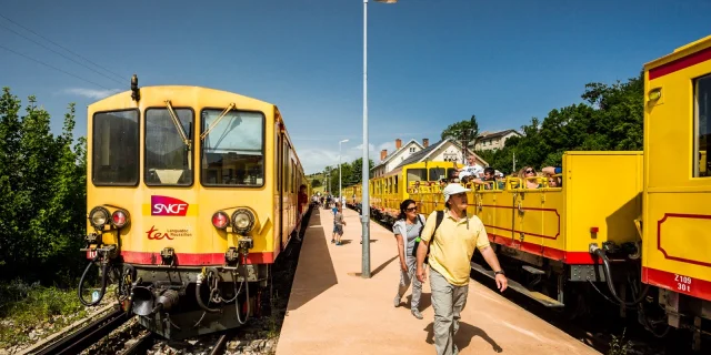 Le Train Jaune à quai en gare de Mont-Louis, avec des voyageurs descendant du train ou montant à bord. Les wagons jaunes emblématiques sont visibles sous un ciel bleu dégagé.