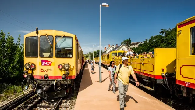 Le Train Jaune à quai en gare de Mont-Louis, avec des voyageurs descendant du train ou montant à bord. Les wagons jaunes emblématiques sont visibles sous un ciel bleu dégagé.