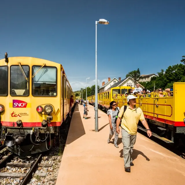 Le Train Jaune à quai en gare de Mont-Louis, avec des voyageurs descendant du train ou montant à bord. Les wagons jaunes emblématiques sont visibles sous un ciel bleu dégagé.