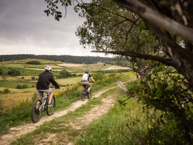 Deux personnes en VTT électrique roulant sur un sentier herbeux avec un paysage vallonné et verdoyant en arrière-plan.