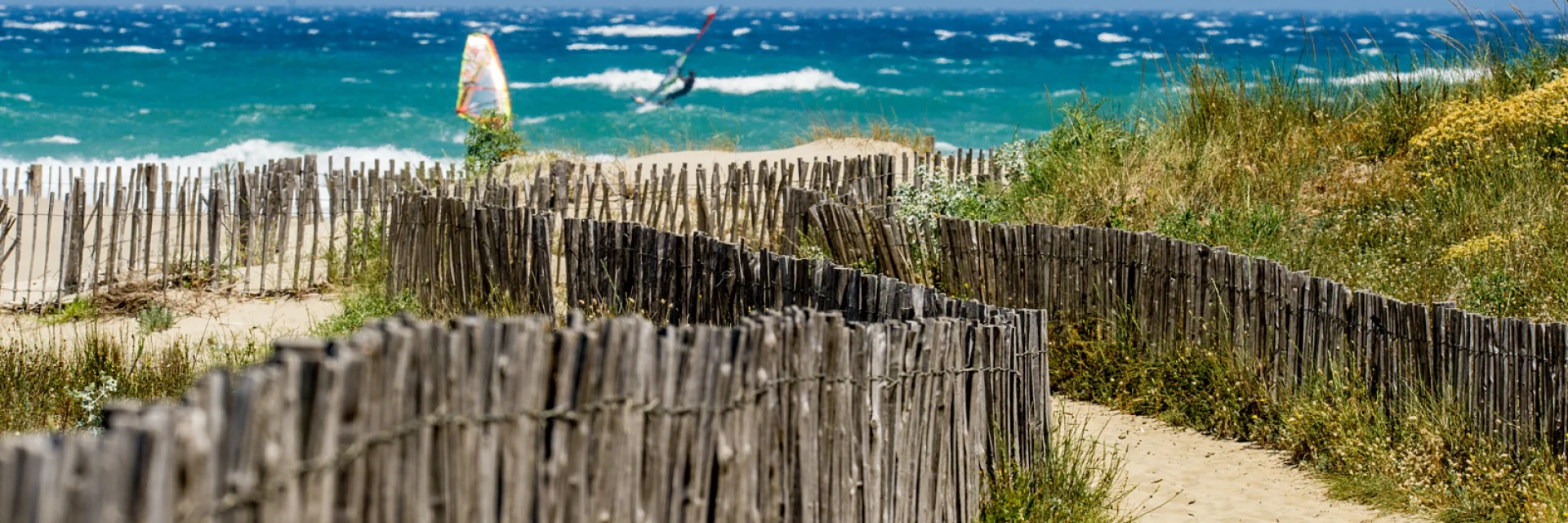 Sentier sableux parmi des ganivelles menant à une plage venteuse où des planchistes naviguent sur des vagues.