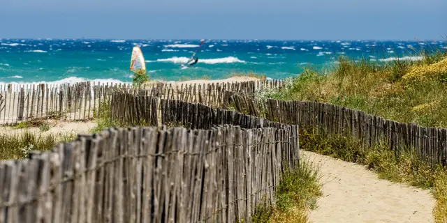 Sentier sableux parmi des ganivelles menant à une plage venteuse où des planchistes naviguent sur des vagues.