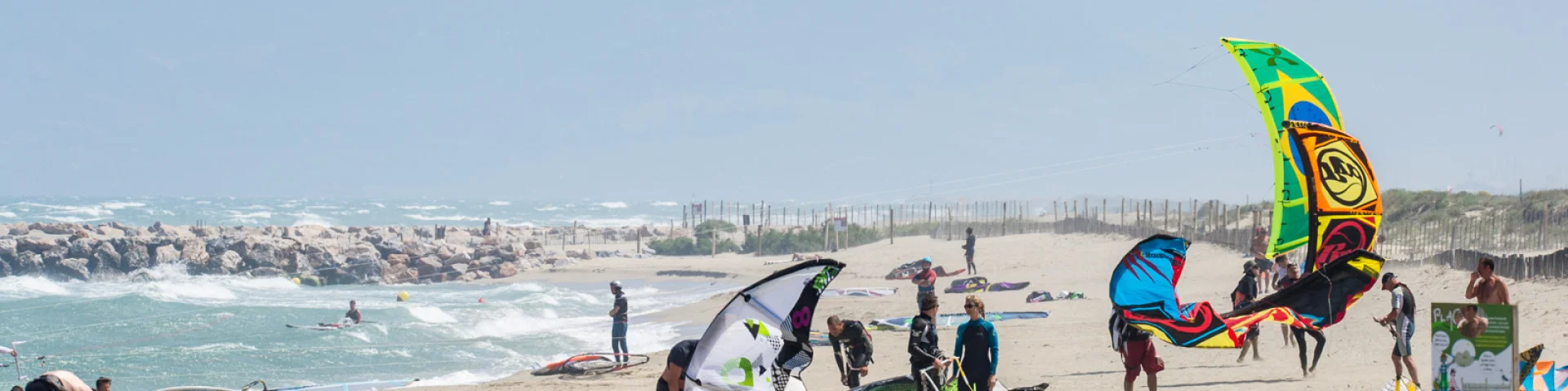 Plage ventée avec pratiquants installant leurs ailes de kitesurf près du rivage.
