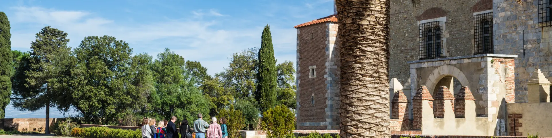 Groupe de visiteurs dans une cour ensoleillée près d’un grand bâtiment en pierre et d’un palmier.