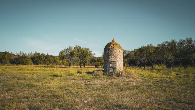 Petite construction en pierre, de forme cylindrique avec un toit arrondi, isolée au milieu d’un champ herbeux entouré d’arbres.