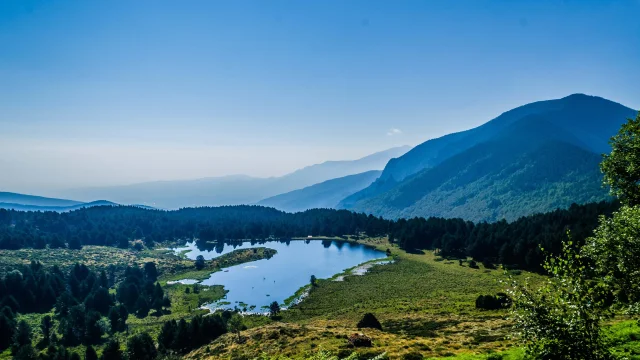 Paysage de montagne avec un lac au centre, entouré de prairies et de forêts, sous un ciel bleu dégagé.