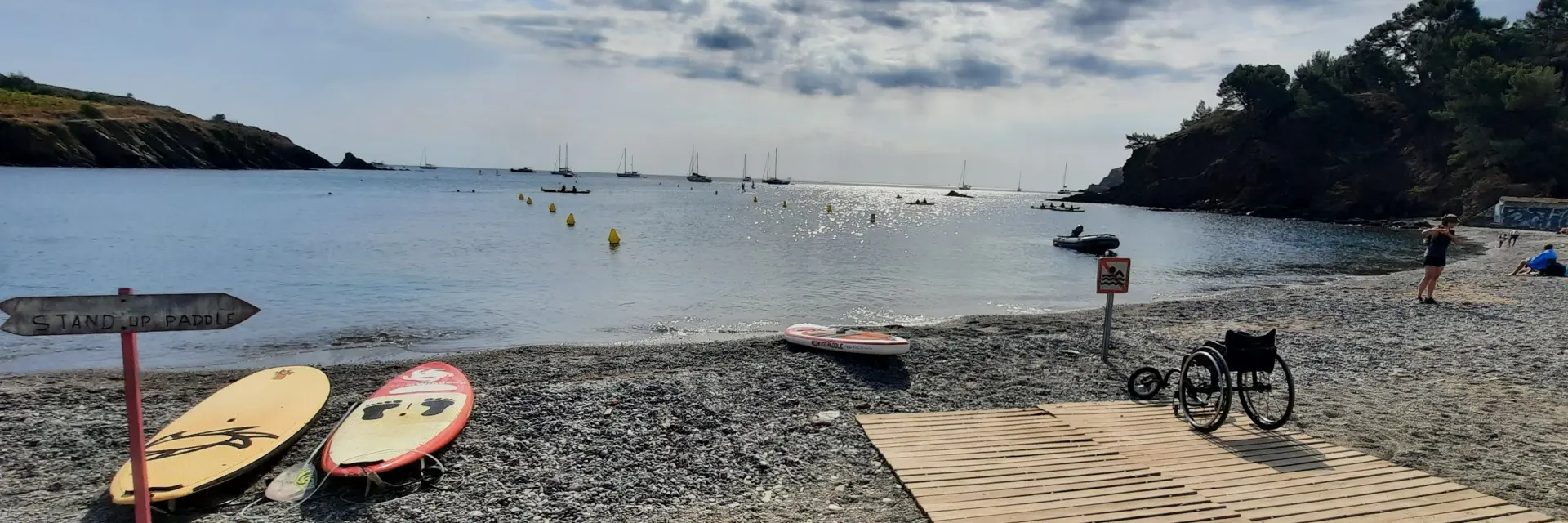 Plage de galets avec rampe d’accès en bois menant à la mer, équipée pour les personnes à mobilité réduite. Planche de stand up paddle et fauteuil roulant visibles au premier plan, mer calme et voiliers à l’horizon sous un ciel partiellement nuageux.