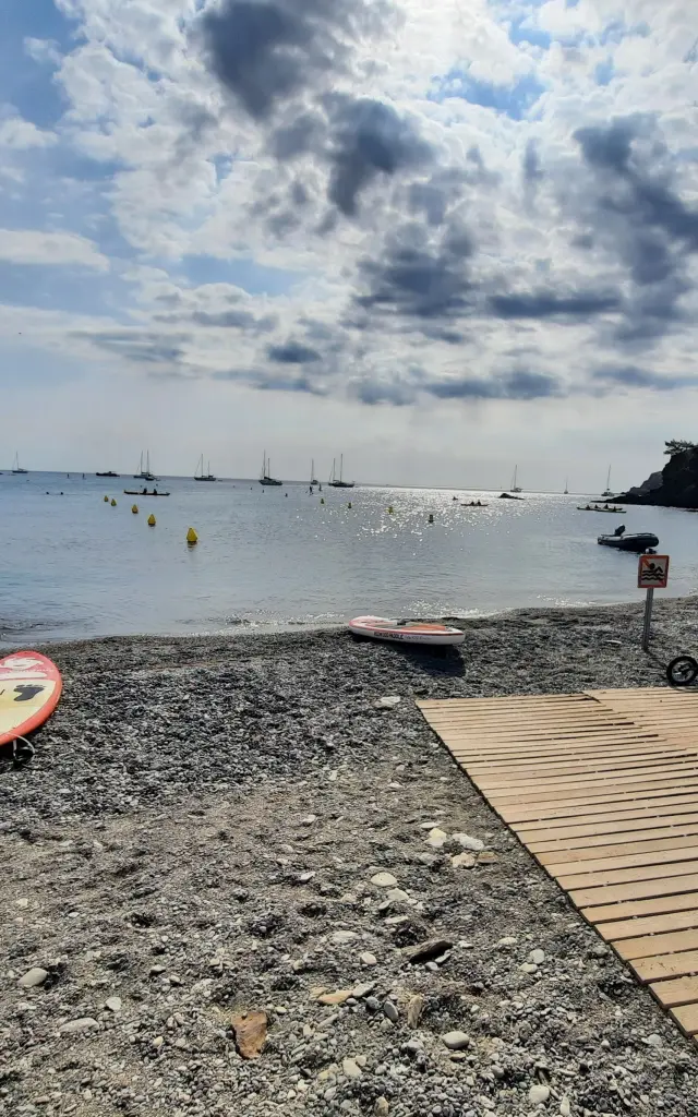 Plage de galets avec rampe d’accès en bois menant à la mer, équipée pour les personnes à mobilité réduite. Planche de stand up paddle et fauteuil roulant visibles au premier plan, mer calme et voiliers à l’horizon sous un ciel partiellement nuageux.