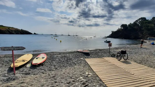 Plage de galets avec rampe d’accès en bois menant à la mer, équipée pour les personnes à mobilité réduite. Planche de stand up paddle et fauteuil roulant visibles au premier plan, mer calme et voiliers à l’horizon sous un ciel partiellement nuageux.