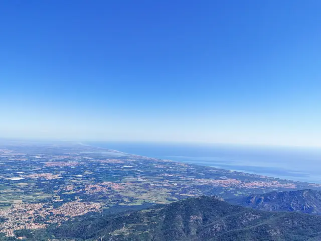 Panorama depuis un sommet sur une plaine cultivée, des villages et la côte méditerranéenne sous un ciel bleu.
