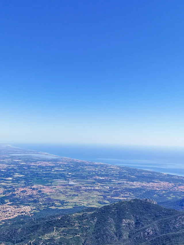 Panorama depuis un sommet sur une plaine cultivée, des villages et la côte méditerranéenne sous un ciel bleu.