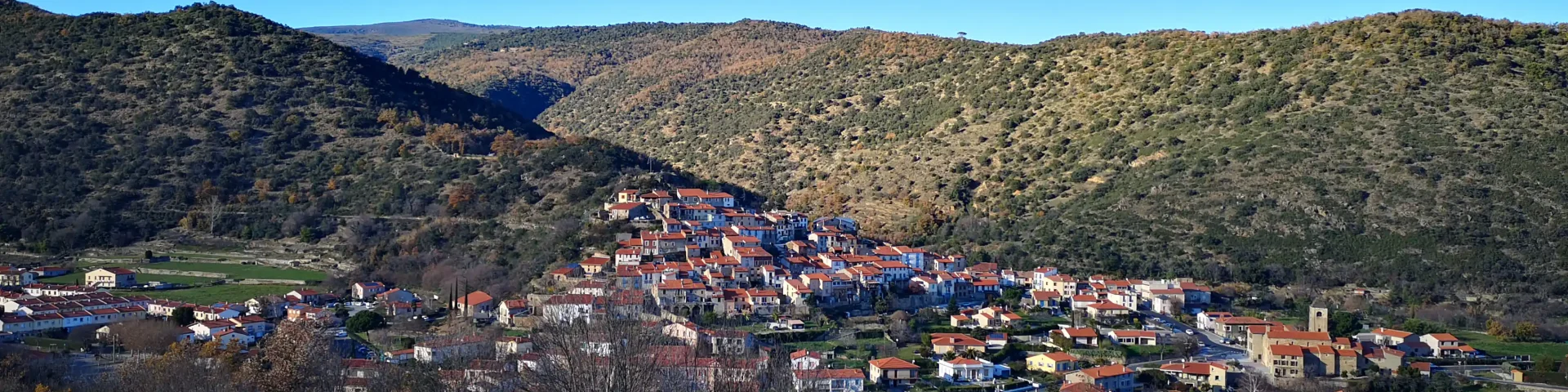 Vue d’ensemble d’un village aux toits ocres installé sur une pente, entouré de collines sèches et d’un ciel bleu dégagé.